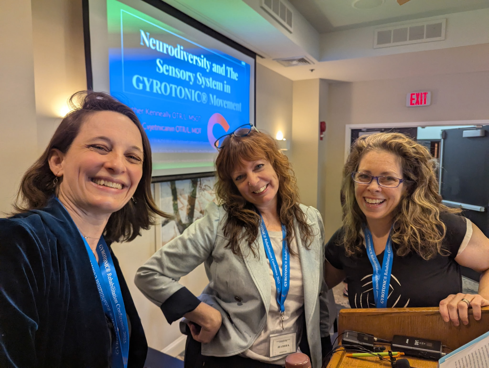 Three women Gyrotonic trainers standing in front of a presentation screen that reads "Neurodiversity and the Sensory System in GYROTONIC® Movement"